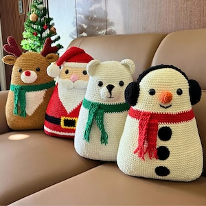 May include: Four handmade crochet Christmas pillows in the shape of a reindeer, Santa Claus, polar bear, and snowman. The reindeer is brown with antlers and a green scarf. Santa is red with a white beard. The polar bear is white with a green scarf. The snowman is white with a red scarf.