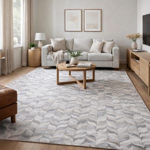 May include: A large area rug with a wave pattern in shades of gray and white dominates the living room. A light gray sofa with beige pillows and a wooden coffee table are in the center. The room has a neutral color palette with natural light.