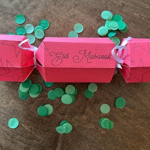 May include: A red paper cracker with a white ribbon and the words "Eid Mubarak" written on it. Green confetti is scattered around the cracker.