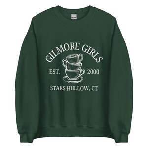 May include: A dark green sweatshirt with the text "GILMORE GIRLS" above a stack of three teacups, with "EST. 2000" and "STARS HOLLOW, CT" below. The design is in white, and the sweatshirt has a crew neck.