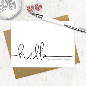 May include: A white card with the word "hello" written in cursive with a black pen. The text "from savannah matthews" is written below the word "hello".