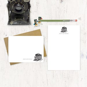 May include: Two white note cards with a black and white illustration of an antique camera. The note cards have the text "Janet Goodman" printed on them.