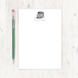 May include: A white notepad with a black and white illustration of an antique camera. The notepad has the text "janet_goodman" printed in gray at the bottom.