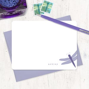 May include: A white card with a purple dragonfly design and the name "ashley" printed at the bottom. The card is on a white wooden surface with a purple envelope and a purple pen.