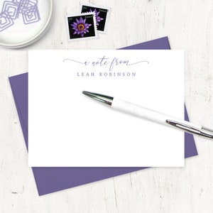 May include: A white note card with a purple envelope. The note card has the text "a note from LEAH ROBINSON" printed on it. A white pen is lying on top of the note card.