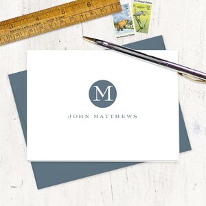 May include: A white card with a blue circle containing the letter "M" in white. The text "JOHN MATTHEWS" is printed below the circle in a dark gray font.