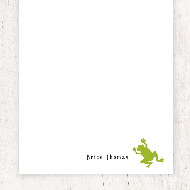 May include: White stationery with the name "Brice Thomas" printed in black. A green frog silhouette is in the lower right corner. The stationery is on a white wooden surface.