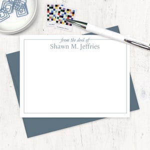 May include: A white stationery set with a blue border, featuring the text "from the desk of Shawn M. Jeffries." A white pen rests on the stationery. Two colorful stamps and a blue envelope are also present.