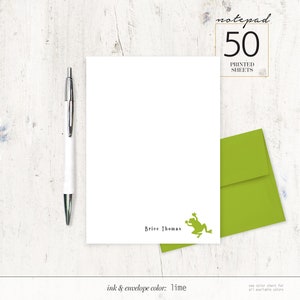 May include: White notepad with the text "Brice Thomas" and a lime green frog illustration. A lime green envelope is next to the notepad. A silver and white pen is on the left. The notepad reads "notepad 50 printed sheets".