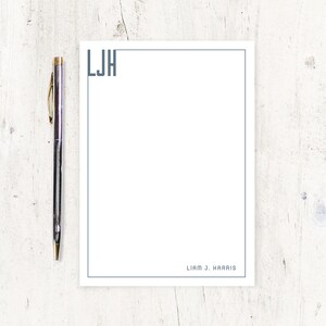 May include: A white notepad with a teal border and the initials "LJH" in the upper left corner. The name "Liam J. Harris" is printed at the bottom. A silver pen with gold accents rests on the left side.