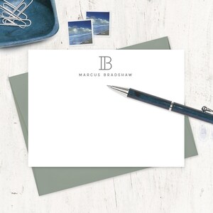 May include: A white personalized stationery set with a gray envelope. The stationery features the initials "IB" above the name "Marcus Bradshaw" in a simple font. A pen with a blue and silver design rests on the stationery, with two stamps and paperclips in the background.