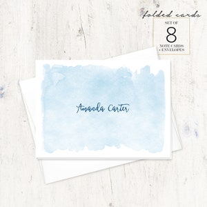 Puede incluir: Un juego de 8 tarjetas plegadas con sobres. Las tarjetas presentan un fondo de acuarela azul claro con el nombre "Amanda Carter" escrito en cursiva azul oscuro.