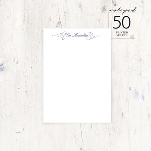 May include: A white notepad with a purple cursive script that says "Ellie Hamilton" at the top. The notepad has a white background and a black border. The notepad is on a white wooden surface. The text "notepad 50 printed sheets" is in a black rectangle in the upper right corner of the image.