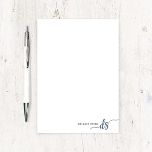 May include: A white notepad with a blue pen on a white wooden surface. The notepad has the text "Delaney Smith" and the initials "ds" in a cursive font.