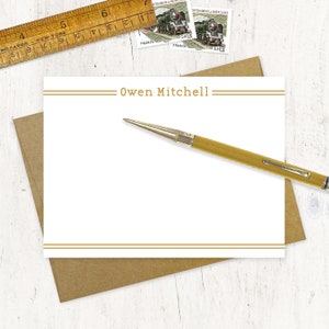 May include: A white stationery card with two thin gold lines at the top and bottom. The name "Owen Mitchell" is printed in gold letters in the center of the card.