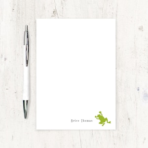 May include: White notepad with a green frog graphic and the name "Brice Thomas" printed in black. A silver and white pen is positioned to the left of the notepad. The notepad is on a white wooden surface.