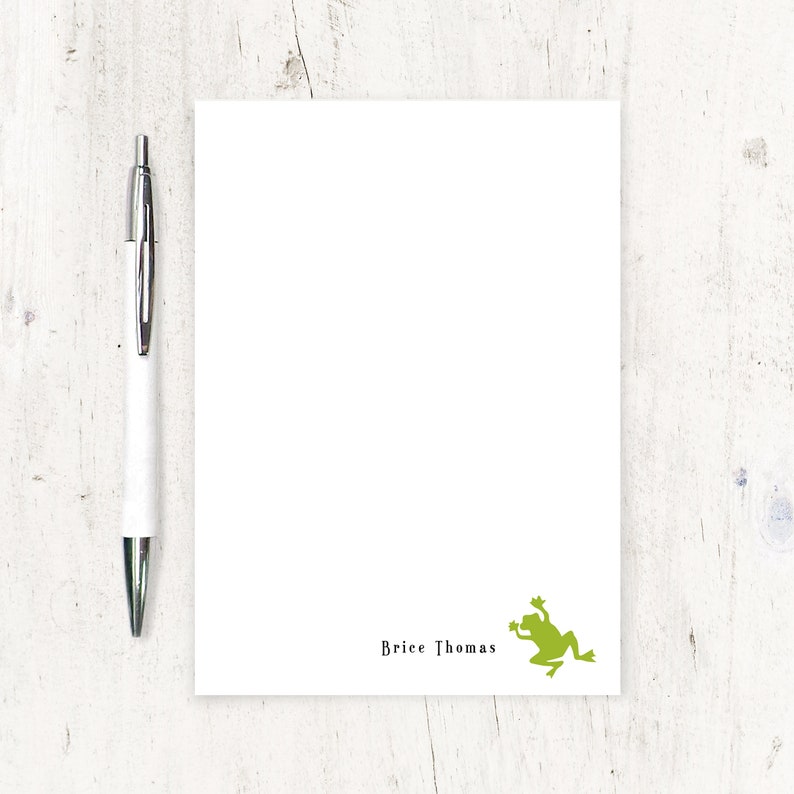 May include: White notepad with a green frog graphic and the name "Brice Thomas" printed in black. A silver and white pen is positioned to the left of the notepad. The notepad is on a white wooden surface.