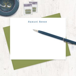 May include: A white card with the name "Samuel Reese" printed in blue text. A blue pen is lying on top of the card. The card is on top of a green envelope.