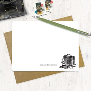 May include: A white card with a black and white illustration of an antique camera. The card has the text "Janet goodman" printed on it.
