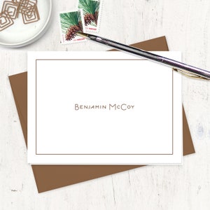 May include: A white card with a brown border and the text "BENJAMIN MCCOY" printed in brown. The card is on a brown envelope and a white wooden surface.