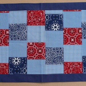 May include: A blue and red patchwork table runner with a blue border. The runner is made of squares of different fabrics, including blue and white gingham, red and white paisley, and solid blue.