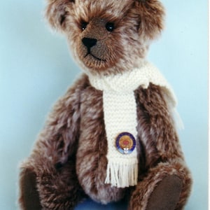 May include: A brown teddy bear wearing a white knitted scarf with a blue and gold pin. The bear has a brown nose and black eyes.