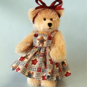 Könnte beinhalten: Ein Plüschteddybär, der eine rote Schleife und ein kariertes Kleid mit Lebkuchen-Designs trägt. Das Kleid ist rot-blau-weiß kariert mit Lebkuchen und Sternen.