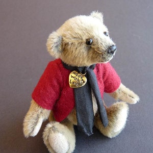 Peut inclure: Un petit ours en peluche beige portant une veste rouge et une cravate noire. L'ours a une étiquette en forme de cœur qui dit "Made with love".