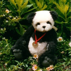 Könnte beinhalten: Ein schwarz-weißes Plüschpanda-Spielzeug, das ein rotes Band mit einer goldenen Medaille trägt. Der Bär sitzt in einem Bett aus grünen Pflanzen mit weißen Blüten.
