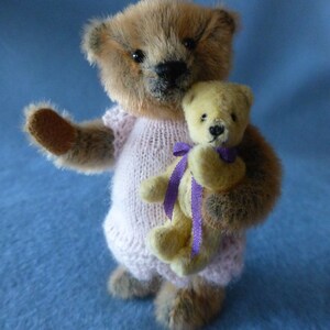 May include: A brown teddy bear wearing a pink knitted sweater is holding a smaller yellow teddy bear with a purple ribbon.