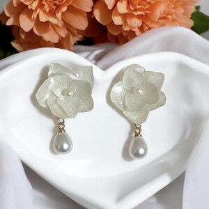Peut inclure: Une paire de boucles d'oreilles en forme de fleurs blanches, avec une perle au centre et un pendentif en forme de goutte de perle. Les boucles d'oreilles sont fixées sur du métal doré. Elles sont présentées sur un plat blanc en forme de cœur.