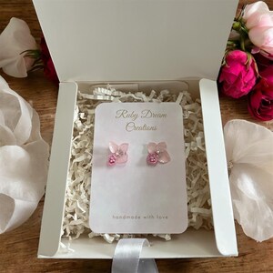 Peut inclure: Boucles d'oreilles clous roses en forme de fleurs, présentées dans un coffret cadeau blanc. Les boucles d'oreilles sont sur une carte blanche avec le texte "Ruby Dream Creations" et "handmade with love". La boîte est remplie de papier déchiqueté blanc et décorée de roses roses.