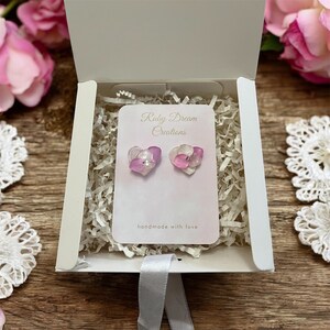 Peut inclure: Boucles d'oreilles en forme de cœur avec un motif floral, présentées dans une boîte cadeau blanche. Les boucles d'oreilles sont roses et blanches avec un petit cristal au centre. La boîte contient du papier de rembourrage blanc et une carte rose avec "Ruby Dream Creations".