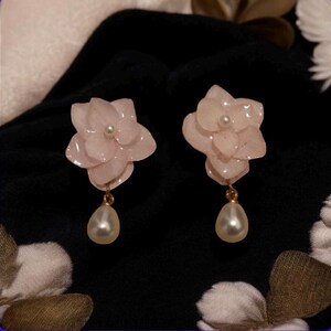 Peut inclure: Une paire de boucles d'oreilles en forme de fleurs rose clair. Chaque boucle d'oreille présente une petite perle au centre de la fleur et un pendentif en forme de goutte de perle suspendu à un fil doré. Les boucles d'oreilles sont posées sur un fond noir.