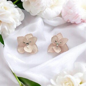 Peut inclure: Deux boucles d'oreilles en forme de fleur rose pâle, ornées d'un bijou argenté au centre, posées sur un tissu de satin blanc. Des pivoines blanches et roses entourent les boucles d'oreilles, pour une ambiance romantique.