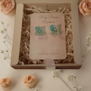 Peut inclure: Une paire de boucles d'oreilles en forme de fleur bleu clair avec des accents de paillettes et un centre de perle, présentée dans une boîte cadeau. La carte indique "Ruby Dream Creations" et "handmade with love."
