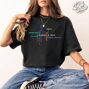 May include: Black t-shirt with a Formula One racing theme. The shirt features the text "FORMULA ONE" and team names like "MERCEDES" and "RACING BULLS" in various colors. The shirt is worn with blue jeans.