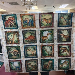 May include: A large quilt featuring a repeating pattern of dinosaur illustrations within square frames. The dinosaurs are depicted in various colors and poses, set against a backdrop of lush greenery. The quilt's border has a floral design.