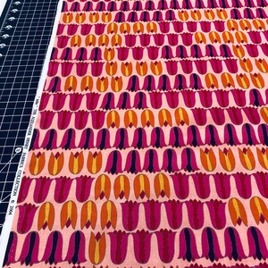 May include: Pink fabric with a repeating pattern of stylized orange, yellow, and pink tulips. The fabric is labeled "The Alexander Henry Fabrics Collection © 2004"