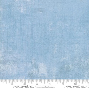 May include: A square of light blue fabric with a textured, distressed appearance. The fabric has a mottled effect with lighter areas and vertical streaks. A ruler is at the bottom, with the word "moda" and website address.