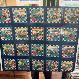 May include: A large, rectangular fabric featuring a repeating pattern of cartoon race cars driven by various animals. Each square has a blue border with a checkered flag design. The cars are red, blue, and yellow.