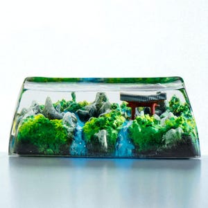 Könnte beinhalten: Ein rechteckiger, klarer Resin-Keycap mit einer Miniaturlandschaft. Die Szene umfasst grünes Laub, graue Felsformationen, einen blauen Wasserfall und ein kleines rotes Torii. Der Keycap ist vor einem weißen Hintergrund platziert.