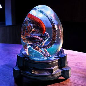 Puede incluir: Una esfera de vidrio transparente con una criatura alienígena detallada en su interior. La criatura está enroscada en posición fetal con una cola larga. La esfera está sobre una base negra con una superficie texturizada.