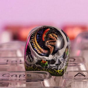 Puede incluir: Una tecla de teclado personalizada con un diseño detallado de calavera, una serpiente y una vibrante paleta de colores. La tecla se encuentra sobre un fondo de teclas de teclado transparentes, con la palabra "Caps" visible en una tecla.