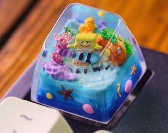 May include: A custom keycap featuring a miniature underwater scene with SpongeBob SquarePants. The keycap is clear with a blue and turquoise base, coral, and other sea creatures. The keycap is square and sits on a keyboard.