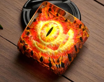 May include: A square, clear glass paperweight with a red, orange, and yellow eye design. The eye is surrounded by a black and white pattern.