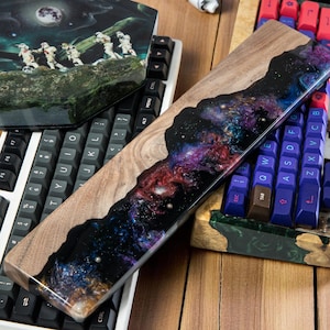 Könnte beinhalten: Eine Holz-Handgelenkauflage für eine Tastatur mit einem Galaxie-Epoxy-Harzeinlage. Das Harz ist eine Mischung aus blauen, violetten und rosa Farben mit schwarzen Wirbeln. Die Handgelenkauflage liegt auf einer weißen Tastatur mit schwarzen Tasten.
