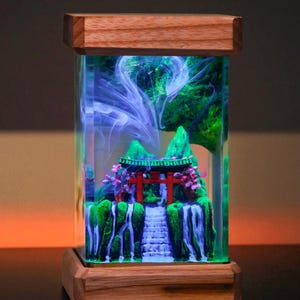 Könnte beinhalten: Rechteckige Kunstharzlampe mit einer japanischen Miniaturlandschaft. Die Szene umfasst ein rotes Torii-Tor, Wasserfälle und grünes Laub. Die Lampe hat einen Holzsockel und -deckel, und der Innenraum hat wirbelnde blaue und weiße Akzente.