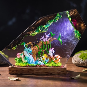 May include: A resin diorama featuring multiple small Eevee evolution figures arranged on a mossy base with a wooden stand. The scene includes a central tree branch, green foliage, and a translucent resin backdrop with purple lighting effects.