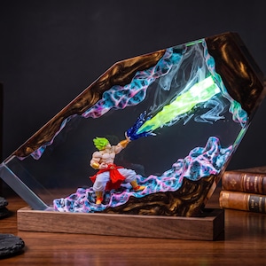 May include: A decorative resin lamp featuring a sculpted figure with spiky green hair, wearing white trousers and a red sash, emitting a bright green energy blast. The figure is encased in a clear, angular resin block with dark wood accents.